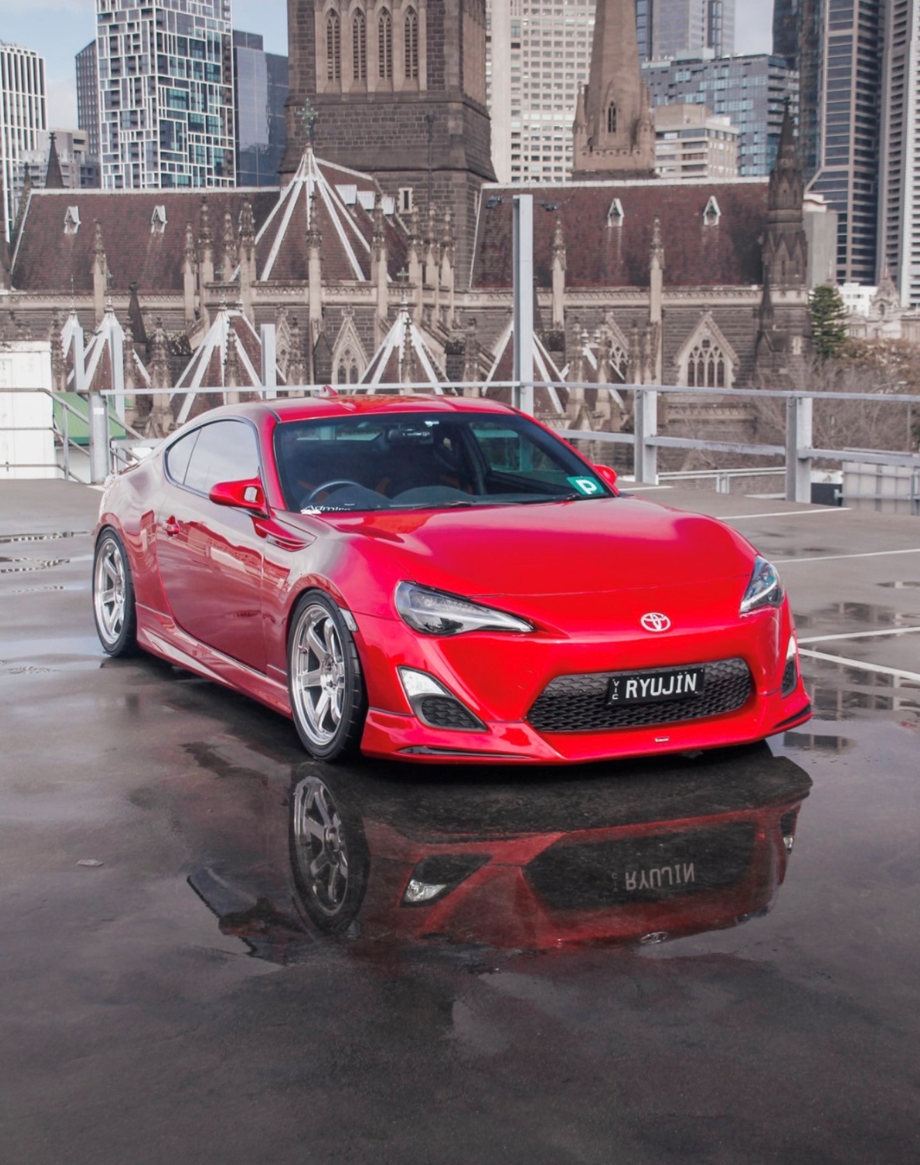 Red Toyota 86 on a wet Melbourne rooftop car park with St Patrick's Cathedral and the CBD behind it