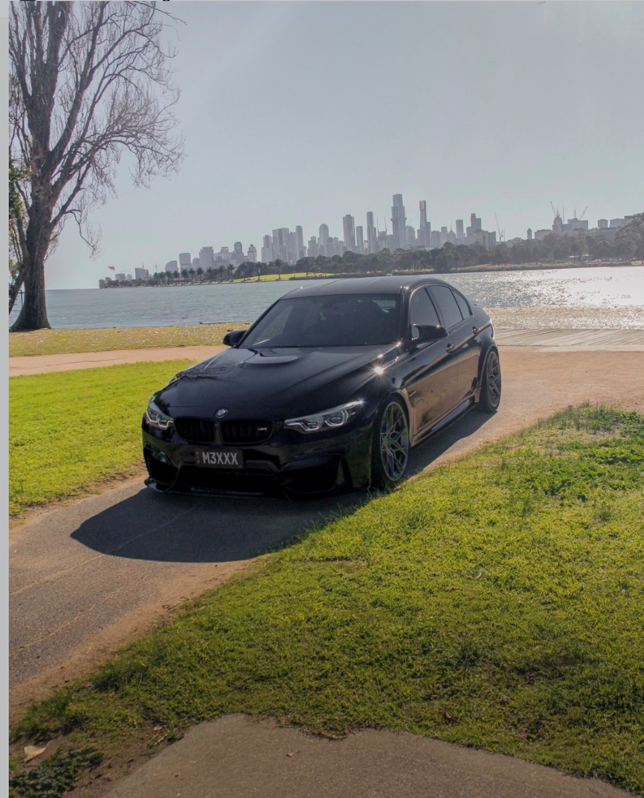 Black BMW M3 parked by Albert Park lake with the Melbourne skyline behind