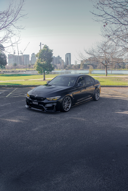 Black BMW M3 sedan parked by Albert Park lake with the Melbourne skyline in the background